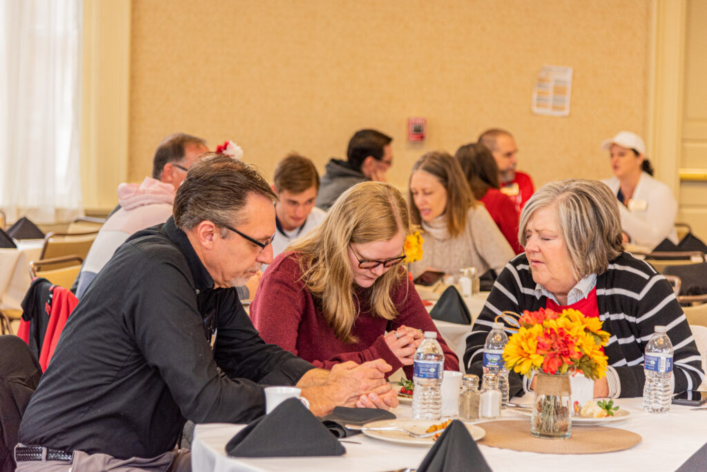 A familiy at a round table in prayer before a meal at a Christian college event, embodying a moment from a real-life prayer guide focused on gratitude, fellowship, and faith in everyday life. This event was called: prayer for college students.