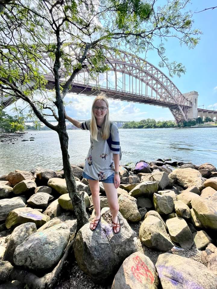 Young woman standing on river rocks near a bridge during a summer internship in NYC, reflecting on creation and living out her faith in everyday adventures.