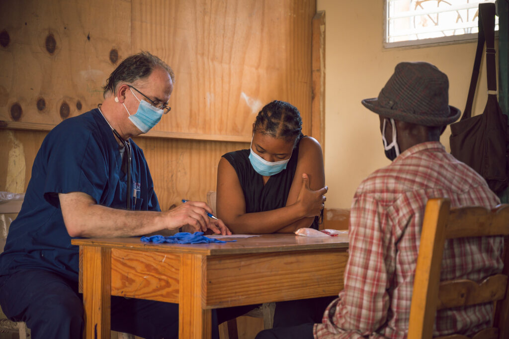 A healthcare professional wearing a mask speaks with two individuals at a wooden table in a modest clinic setting, demonstrating how a Christian worldview informs compassionate care and mission-driven service. A healthcare professional wearing a mask speaks with two individuals at a wooden table in a modest clinic setting, demonstrating how a Christian worldview informs compassionate care and mission-driven service.