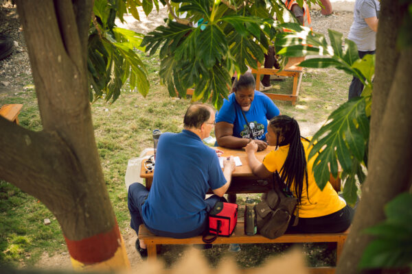 Three people seated at a wooden picnic table under lush trees, engaging in discussion and prayer during a community outreach, reflecting the values of service and compassion shaped by a Christian worldview.