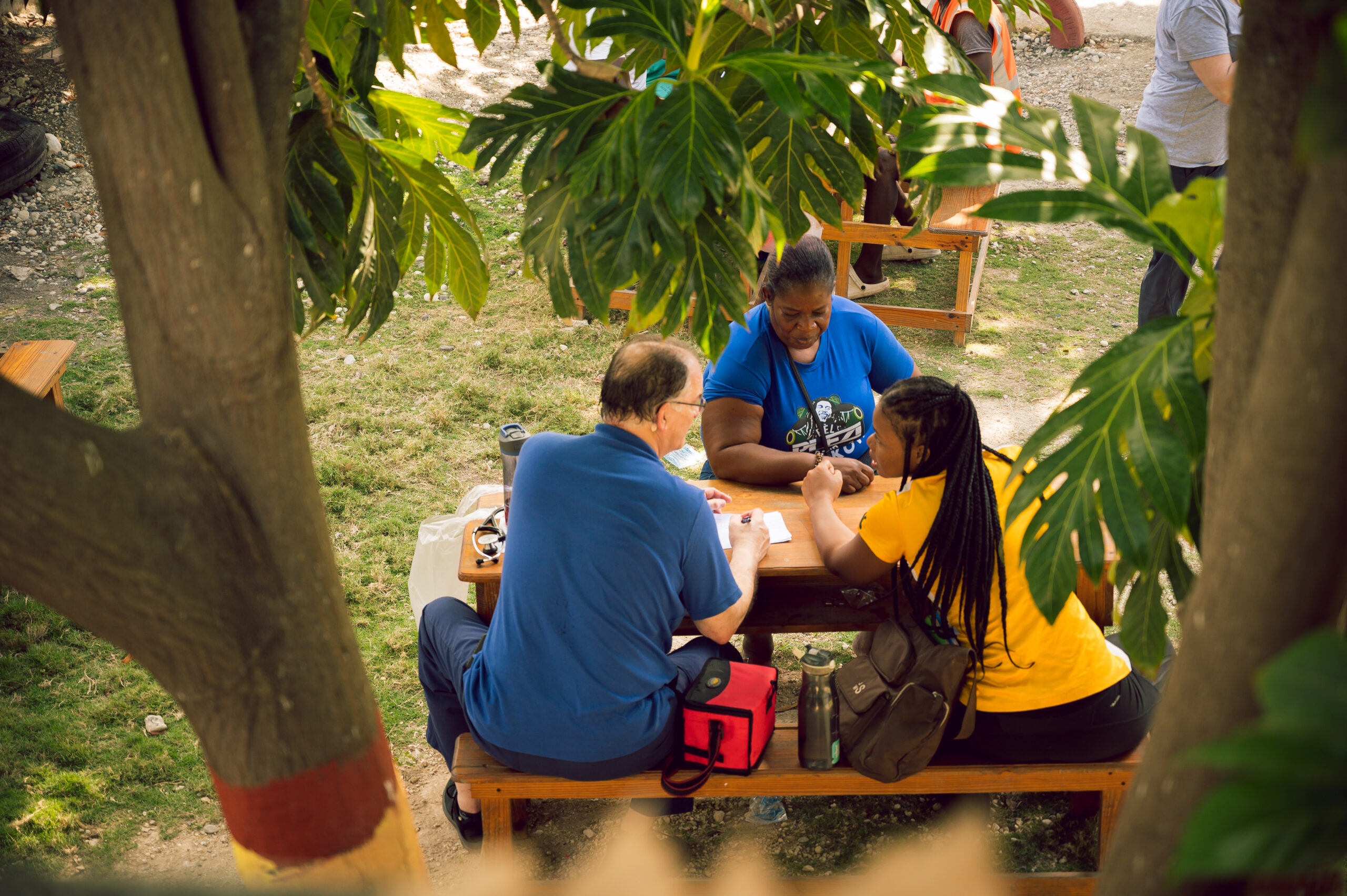 Three people seated at a wooden picnic table under lush trees, engaging in discussion and prayer during a community outreach, reflecting the values of service and compassion shaped by a Christian worldview.