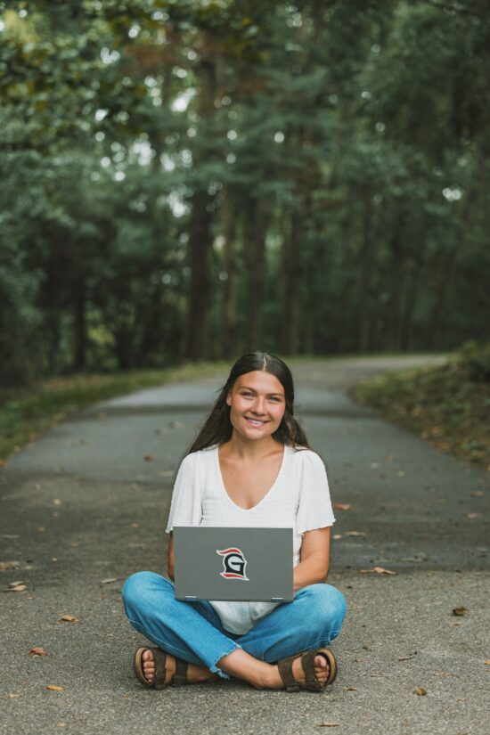 A smiling Grace College student sitting cross-legged on a shaded outdoor path surrounded by trees, representing flexible and accessible online and graduate learning options.