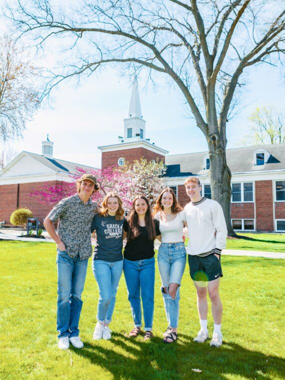 Smiling students standing together on the green lawn in front of a McClain at Grace College a Christian college campus, representing community and faith-based education.