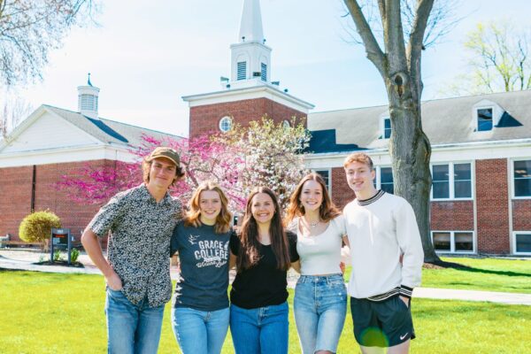 Smiling students standing together on the green lawn in front of a McClain at Grace College a Christian college campus, representing community and faith-based education.