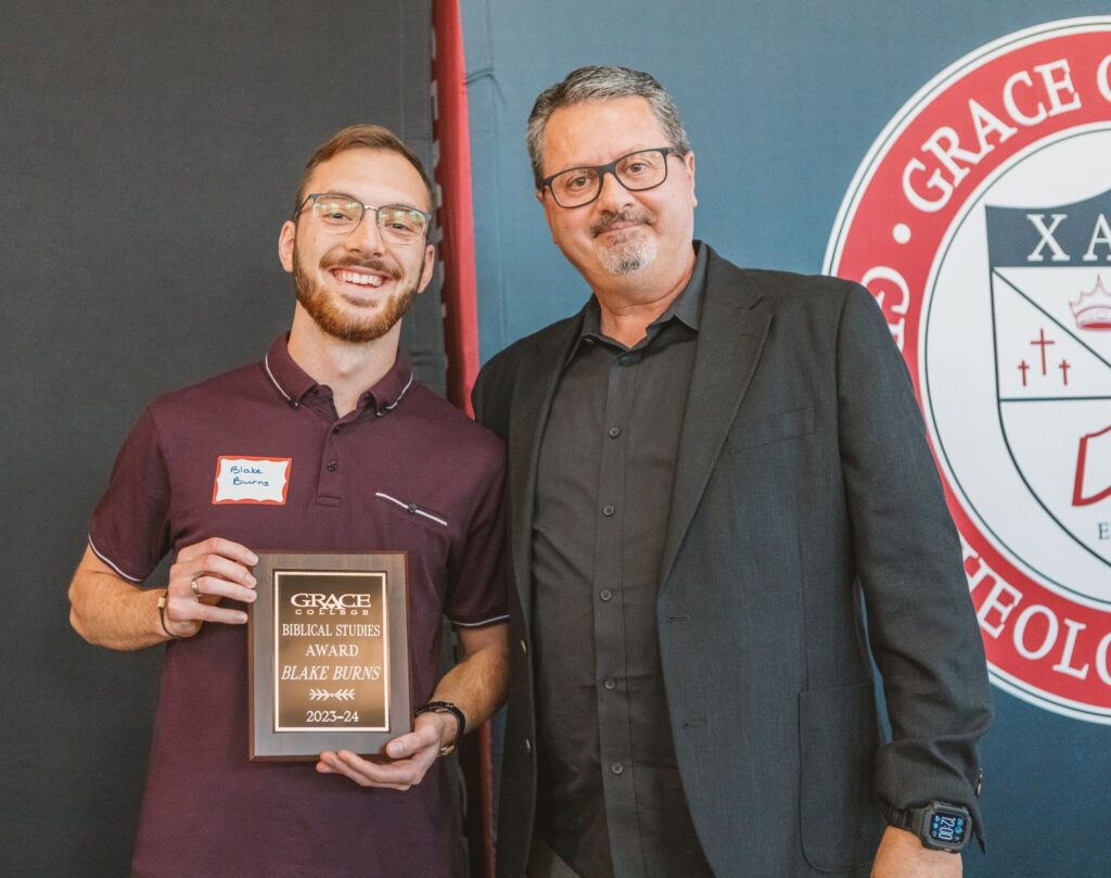 Blake Burns smiles while holding an award plaque next to professor Rata at Grace College, symbolizing recognition through Christian mentorship.