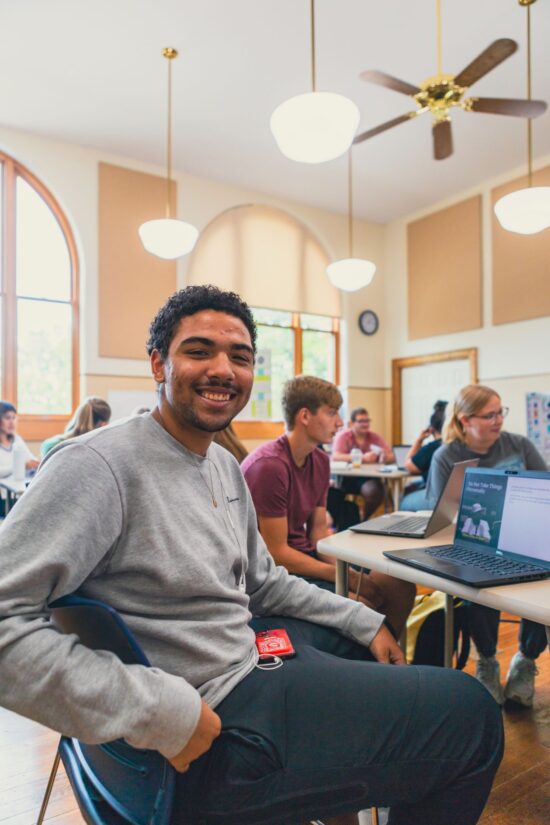A cheerful student in a classroom setting with peers using laptops, showcasing Grace College's opportunities for high school students to earn college credit.